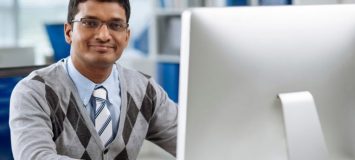 A close up of a happy employee in a sweater vest and tie, looking at the camera from his desk. Get a new career, start your CCIE, Routing and Switching Certificate Course at CCBST.
